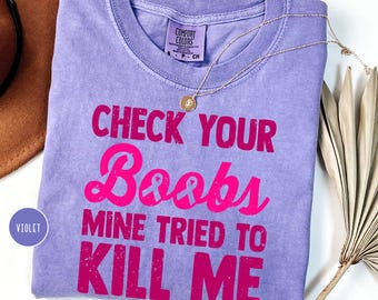 May include: A violet short-sleeved t-shirt with a crew neckline and a Comfort Colors label. The shirt features the hot pink text "CHECK YOUR Boobs MINE TRIED TO KILL ME" with a pink ribbon icon integrated into the word "Boobs".