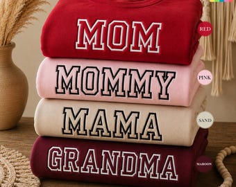 May include: A stack of four crewneck sweatshirts in red, pink, sand, and maroon. Each sweatshirt features a different word in white block letters with a black outline: MOM, MOMMY, MAMA, and GRANDMA. The sweatshirts are folded and stacked on top of each other.