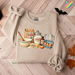 May include: A sand-colored crewneck sweatshirt featuring a graphic print of baking ingredients including a bowl with a wooden spoon, a bag of flour, a jar of sugar, a stick of butter, a bottle of vanilla extract, and a carton of brown eggs.