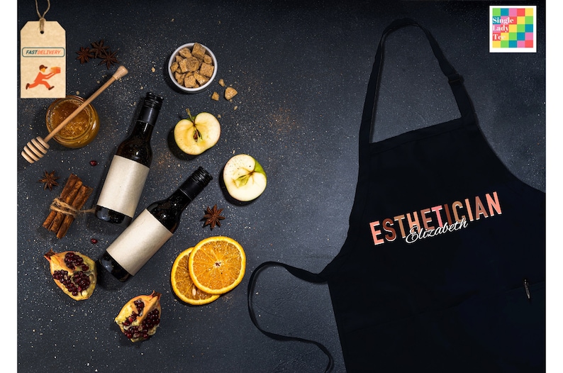 Pode incluir: Avental preto com o texto "ESTHETICIAN Elizabeth" em letras douradas rosadas. O avental &eacute; cercado por v&aacute;rios ingredientes, incluindo ma&ccedil;&atilde;s, laranjas, paus de canela e anis estrelado.