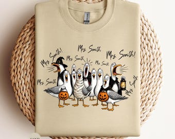 May include: A sand-colored crewneck sweatshirt featuring a graphic of multiple seagulls dressed in various Halloween costumes, including a mummy and a witch, with the text Mrs. Smith repeated several times above and around the birds.