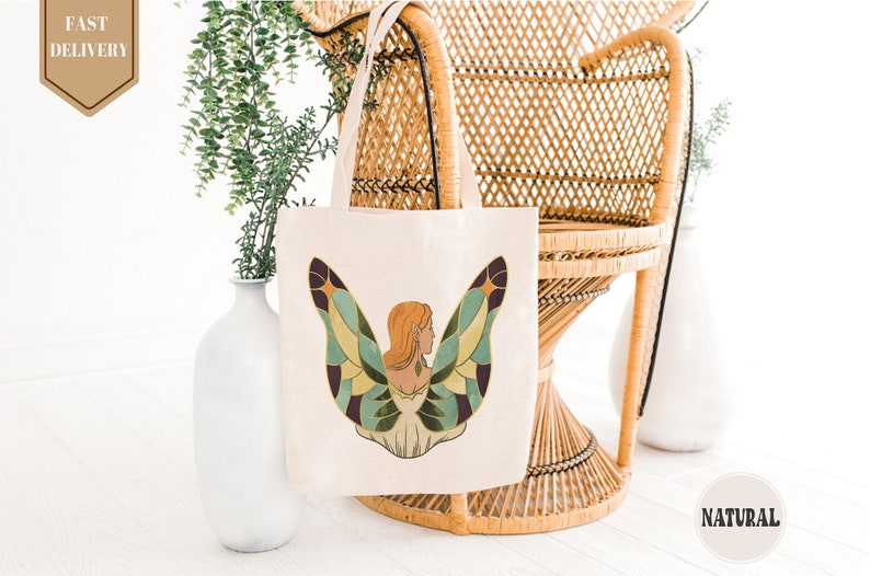 May include: A white tote bag with a colorful illustration of a woman with butterfly wings. The woman has long red hair and is wearing a white dress. The tote bag is sitting on a wicker chair.