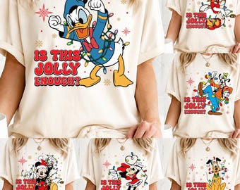 May include: A cream-colored t-shirt featuring a graphic of Donald Duck tangled in colorful Christmas lights with the red text "IS THIS JOLLY ENOUGH?" surrounded by small star and snowflake patterns. The design includes a blue hat and red bow tie.