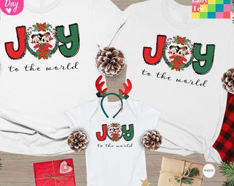 May include: White Christmas-themed t-shirts and a onesie with the word "JOY" in red and green, featuring Mickey and Minnie Mouse. The phrase "to the world" is below the word "JOY". A reindeer antler headband is also included.