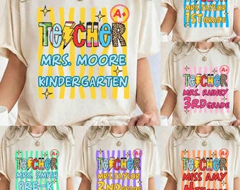 May include: A cream-colored short-sleeve t-shirt featuring a graphic print with yellow vertical stripes. The design includes the text "TEACHER", "MRS. MOORE", and "KINDERGARTEN" in colorful, patterned letters, along with a pencil icon and an A+ symbol.