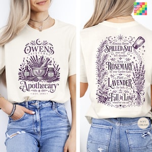 May include: A cream-colored t-shirt featuring a purple graphic print. The front displays a mortar and pestle with the text Owens Sisters Apothecary Est. 1995. The back features botanical illustrations and text about salt, rosemary, and lavender.