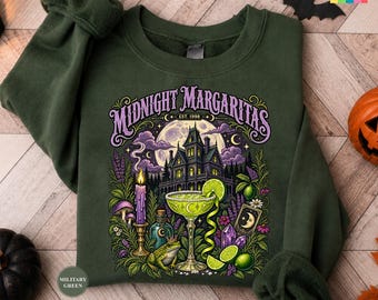 May include: A dark olive green crewneck sweatshirt featuring a graphic print with the text Midnight Margaritas and Est. 1998. The design includes a haunted house, a full moon, a margarita glass, a frog, a purple candle, and lime slices.