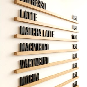 May include: A coffee menu displayed on a white wall. The menu features black lettering spelling out coffee drinks like "Espresso," "Latte," and "Mocha." Each drink name is placed on a light brown shelf. Prices are listed to the right of each drink.