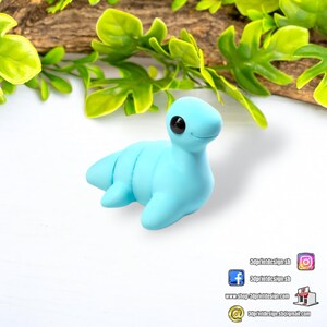 Może przedstawiać: Jasnoniebieska, drukowana w 3D figurka dinozaura z czarnymi oczami. Małe, stylizowane stworzenie leży na białej powierzchni. Zielone liście i kawałek drewna znajdują się w tle. Figurka ma tekst "3dprintdesign.sb" i ikony mediów społecznościowych.