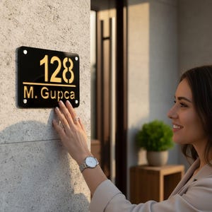Puede incluir: Una placa de dirección rectangular negra con letras doradas. Los números "128" se muestran encima del nombre "M. Gupca". La placa está montada en una pared texturizada de color claro.