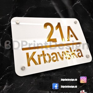 Może przedstawiać: Prostokątny, biały szyld z numerem domu "21A" i nazwą "Krbavska" w kolorze złotym. Szyld jest zamontowany za pomocą czterech srebrnych śrub. Tło to ciemna, marmurowa powierzchnia.