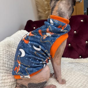 May include: A sleeveless pet sweater with a dark blue fabric featuring a repeating pattern of orange foxes, white crescent moons, and stars. The garment has a bright orange collar and hem, and a small white tag labeled "SEAMSTRESS" at the bottom.