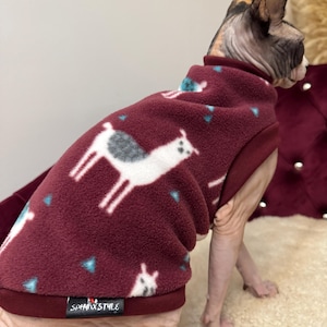 May include: A burgundy fleece pet jumper featuring a repeating white llama pattern with small blue triangular accents. The garment has a high neck, a solid burgundy hem, and a black rectangular label with the white text SPHYNX STYLE.