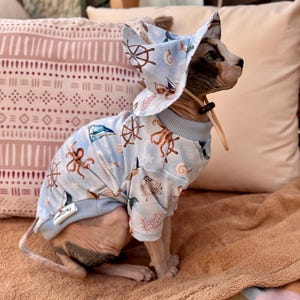 Puede incluir: Una camisa para mascota y un sombrero de pescador a juego en color azul claro con un estampado náutico de pulpos marrones, timones, conchas y coral. La camisa tiene cuello y puños acanalados, y el sombrero incluye una correa ajustable con cierre.
