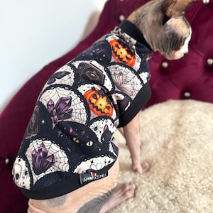 May include: A pet jumper featuring a black and white patterned print with orange jack-o'-lanterns, purple crystals, spiderwebs, and black cat eyes. The garment has a black collar and hem, with a small black tag labelled SPHINX STYLE.