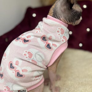 May include: A light pink pet sweater featuring a repeating pattern of white bunny faces wearing heart-shaped sunglasses and pink paw prints. The garment has a high collar, pink trim at the armholes and hem, and a small white label reading SPHYNX STYLE.