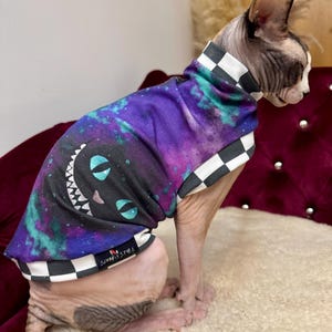 May include: A sleeveless pet shirt featuring a purple and teal galaxy print with a black cat face graphic showing a wide grin and blue eyes. The garment has a black and white chequered trim at the neck and hem, with a small tag reading SPHINX STYLE.
