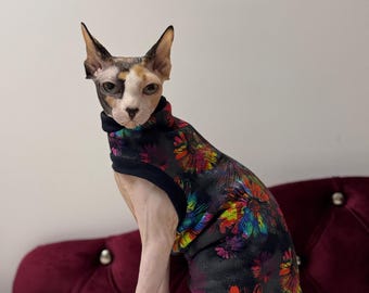 May include: A hairless cat wearing a black and colorful floral print shirt. The cat is sitting on a white fluffy surface with a red velvet couch in the background.