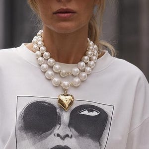 Puede incluir: Una mujer con una camiseta blanca con un gráfico en blanco y negro de una cara. Lleva un collar de perlas en capas con un colgante de corazón dorado, gafas de sol y pantalones grises. Sostiene un bolso de mano negro.