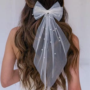 White tulle hair bow with pearl accents. The bow is large and features a cascading tulle veil. The veil is adorned with small, evenly spaced pearls.  The bow is tied in a classic style and is suitable for bridal or special occasion hairstyles. The hair accessory is delicate and elegant.