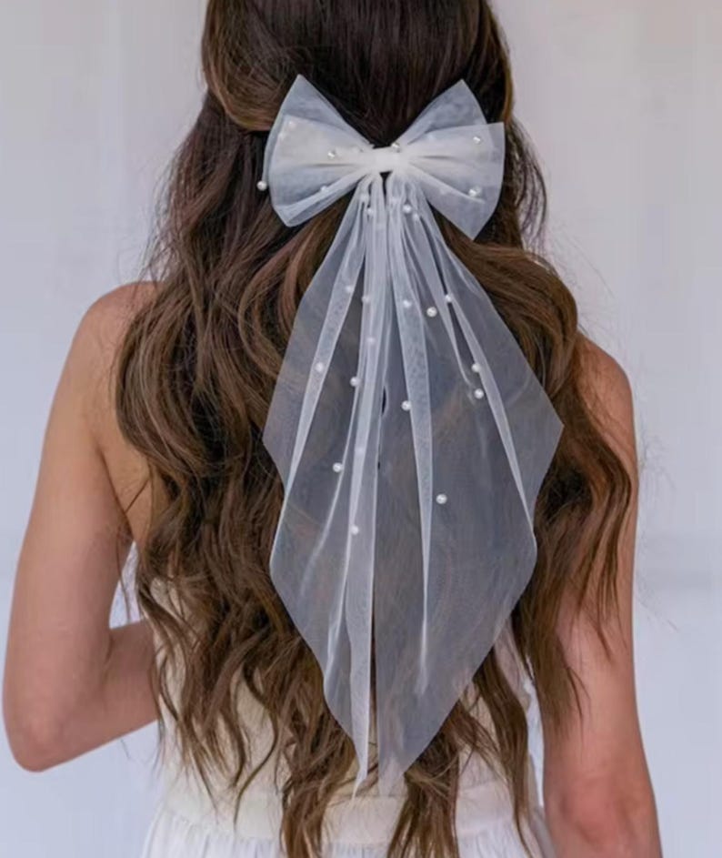 White tulle hair bow with pearl accents. The bow is large and features a cascading tulle veil. The veil is adorned with small, evenly spaced pearls.  The bow is tied in a classic style and is suitable for bridal or special occasion hairstyles. The hair accessory is delicate and elegant.