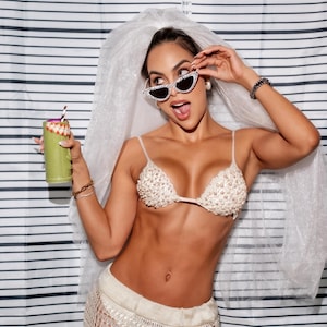 May include: A woman wearing a beaded bikini top, a crochet skirt, and sunglasses, holding a green drink with a straw. The background features a black and white striped pattern with height measurements in inches.