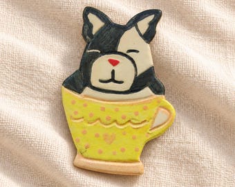 May include: A ceramic brooch shaped like a black and white French Bulldog sitting inside a yellow teacup. The teacup features a scalloped rim, a small handle, and a pattern of light pink polka dots. The dog has a red nose and closed eyes.