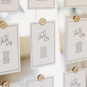 May include: A wedding seating chart display featuring multiple white rectangular cards with black text and borders. Each card is labeled with a table number and a list of names, secured to a surface with circular gold pins embossed with the letters HC.