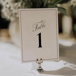 Op de afbeelding: Een tafelnummerkaart met het woord "Table" in sierlijke letters en het nummer "1" in vet zwart. De kaart staat in een zilveren houder, met een wit tafelkleed en bloemstuk op de achtergrond.