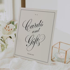May include: A rectangular off-white sign with a thin black border featuring the text "S A" at the top, "Cards and Gifts" in large black cursive script in the center, and "THANK YOU" in small black capital letters at the bottom.