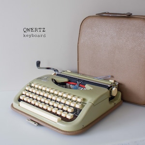 May include: A vintage, pale green typewriter with a QWERTZ keyboard and cream-coloured keys. The typewriter sits on a brown base, next to a matching tan carrying case. The text "QWERTZ keyboard" is visible.