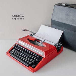 Puede incluir: Una máquina de escribir roja vintage con teclado QWERTZ y maleta de transporte negra. La máquina tiene papel insertado, lista para usar. El texto "QWERTZ keyboard" es visible en el fondo. El modelo de la máquina es BR 200.
