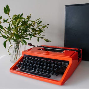 Peut inclure: Une machine à écrire vintage orange vif avec des touches noires. La machine à écrire est posée sur une surface blanche, à côté d'un bocal en verre avec du feuillage vert et un livre noir. Le design de la machine à écrire est rétro.