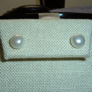 May include: A pair of round, white pearl stud earrings displayed on a textured, light beige fabric earring card. The pearls have a smooth, lustrous surface and are positioned side-by-side on the upper portion of the rectangular display card.