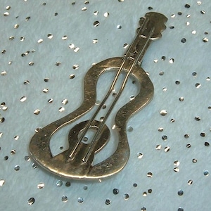 May include: A gold-toned metal hair clip shaped like a guitar. The accessory features a cutout silhouette of the instrument with a long neck, a rounded body, and two parallel bars running down the center to represent strings.