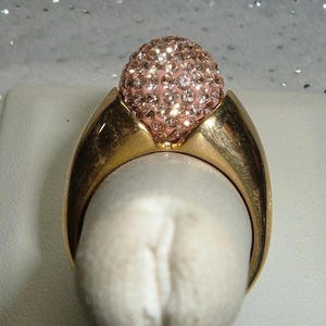 May include: A gold-toned ring with a large, round, pink-toned, crystal-studded top. The ring's band has a smooth, curved design. The ring is set against a white background.