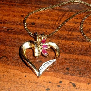 May include: A gold-toned heart pendant necklace with a delicate chain. The heart features a cluster of red gemstones and a section of small white stones. The necklace is displayed on a wooden surface.