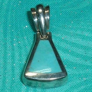 May include: A silver pendant featuring a triangular inlay of smooth, opaque light blue stone. The metal frame has a polished, reflective finish and includes a sturdy, integrated bail at the top for attachment to a chain or cord necklace.