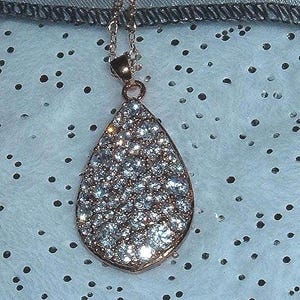 May include: A rose gold teardrop pendant necklace. The pendant is encrusted with numerous small, sparkling clear crystals. The necklace chain is also rose gold. The background is a light blue fabric with black dots.