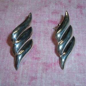 May include: A pair of polished silver-tone earrings featuring a curved, multi-layered wave design. Each earring has a smooth, reflective metallic surface with three distinct, stacked ridges that taper to a point at both the top and bottom ends.