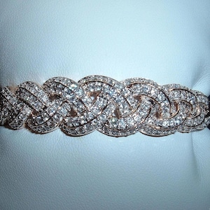May include: A rose gold bracelet with a woven design, encrusted with numerous sparkling, clear gemstones. The bracelet is displayed on a white surface, showcasing its intricate details and luxurious appearance.