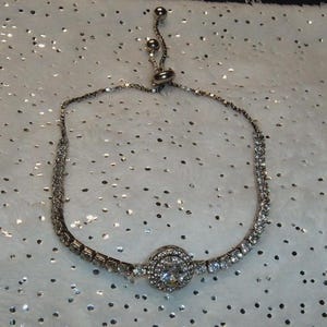 May include: Silver-toned bracelet with a central, round, clear gemstone surrounded by smaller stones. The bracelet features a delicate chain design and an adjustable sliding clasp. The bracelet is set against a white, glittery background.