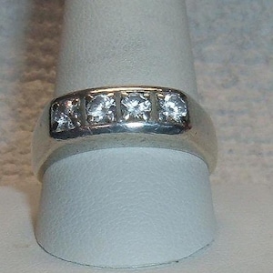 May include: A silver ring with four clear, square-cut stones set in a row. The ring is displayed on a white surface, showcasing its polished metal and sparkling gemstones. The ring's band is smooth and wide.