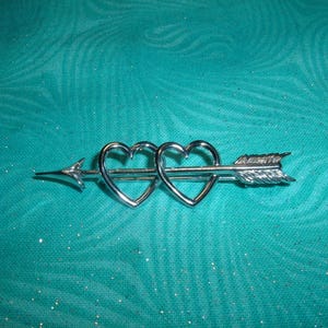 May include: A silver-toned brooch featuring two open-heart shapes pierced by an arrow. The arrow has a pointed tip and feathered fletching. The brooch is set against a teal, textured background.