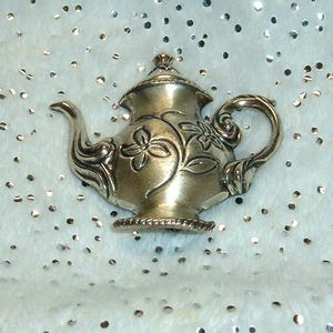 May include: A silver-toned metal teapot brooch featuring an embossed floral pattern on the body, a curved handle, a spout, and a lid. The teapot has a textured base and a polished metallic finish with intricate engraved details on the surface.