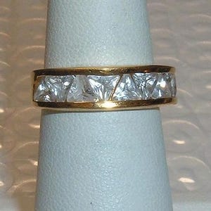 May include: A gold-toned ring with a band of clear, square-cut stones. The ring is set on a white display stand, showcasing the jewelry's design and sparkle. The ring's band is approximately 0.25 inches wide.