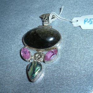 May include: A silver pendant with a black oval stone, three pink stones, and a green and blue abalone shell. The pendant is on a blue fabric background.