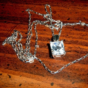 May include: A silver necklace with a square, clear gemstone pendant. The chain is made of small, rectangular links. The necklace is lying on a dark brown, wooden surface, creating a contrast between the shiny metal and the wood.
