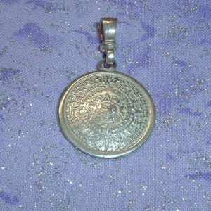 May include: A circular silver pendant featuring an intricate, embossed Aztec calendar design on its face. The pendant includes a small, polished silver bail at the top for attachment to a chain, showcasing a metallic finish and detailed relief.