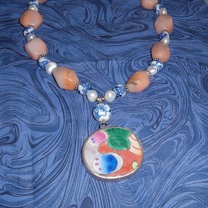 May include: A necklace featuring peach-colored stone beads, white pearls, and blue and white patterned beads. A round pendant with a colorful floral design hangs from the necklace. The necklace is displayed on a blue patterned surface.